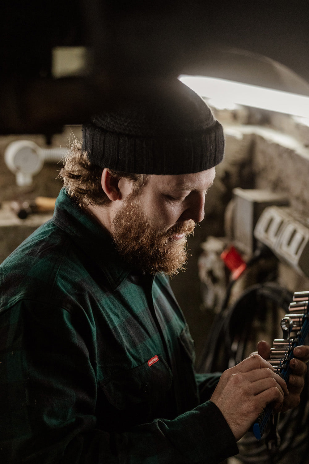 Man with a beard wearing a black beanie and green jacket working on a tool in a workshop.