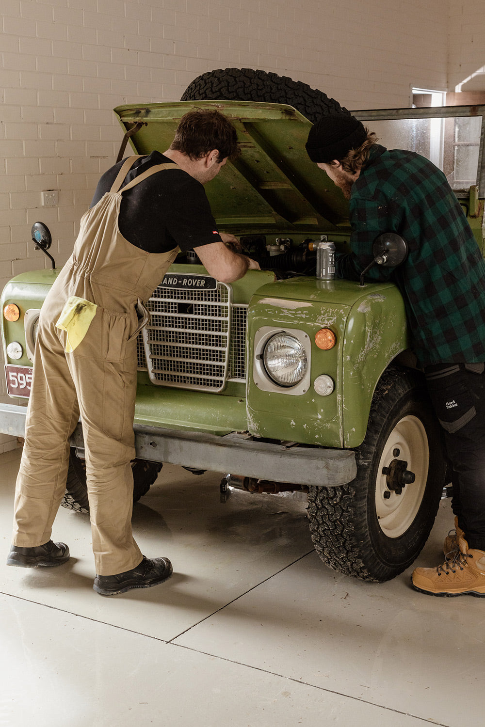 Two people working on a vintage green Land Rover in a garage.