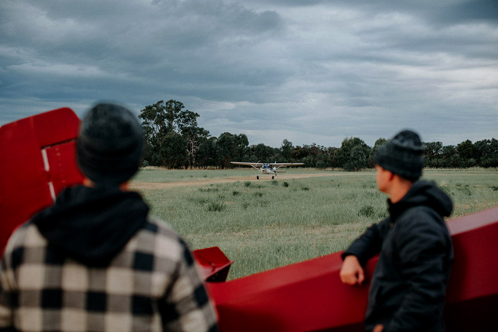 Two people standing near a red airplane on a grassy field with a cloudy sky.