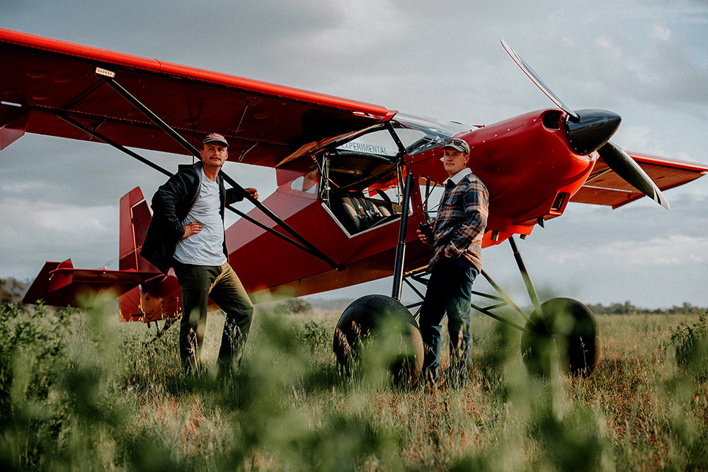 Two men standing next to a red biplane in a grassy field.