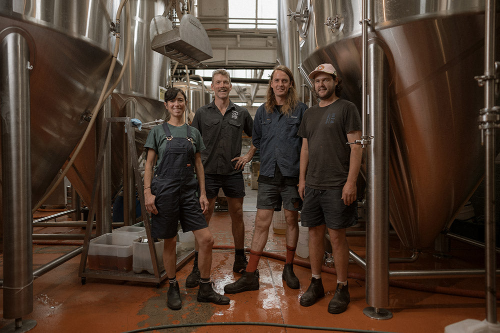 Four people standing in a brewery with large metal tanks and equipment.