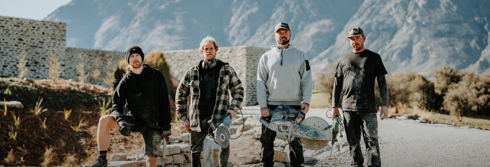 Four men with skateboards standing on a road with mountains in the background