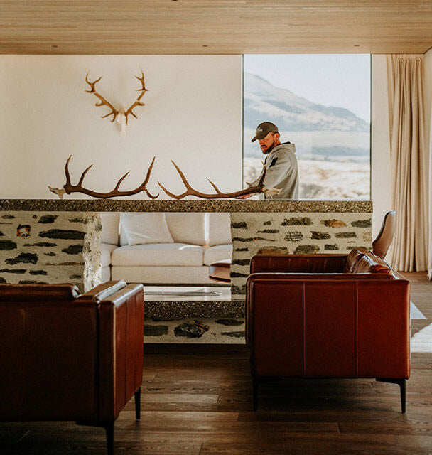 Man standing in a room with stone wall, large window, and deer antlers on the wall.