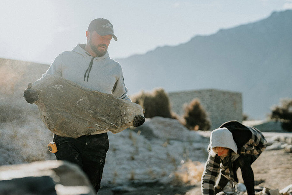 Man holding a large rock in a desert-like setting with mountains in the background
