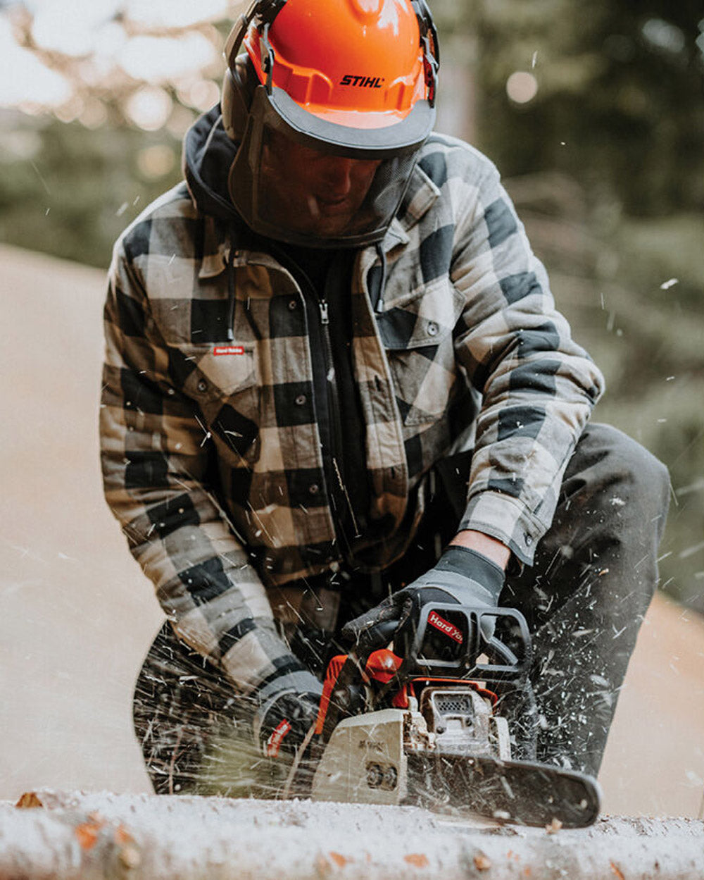 Person using a chainsaw wearing a plaid jacket and orange helmet in a forest setting