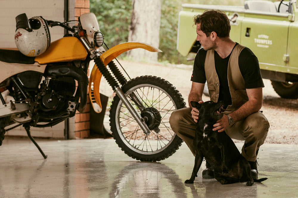Man holding a dog in a garage with a motorcycle and vintage truck in the background