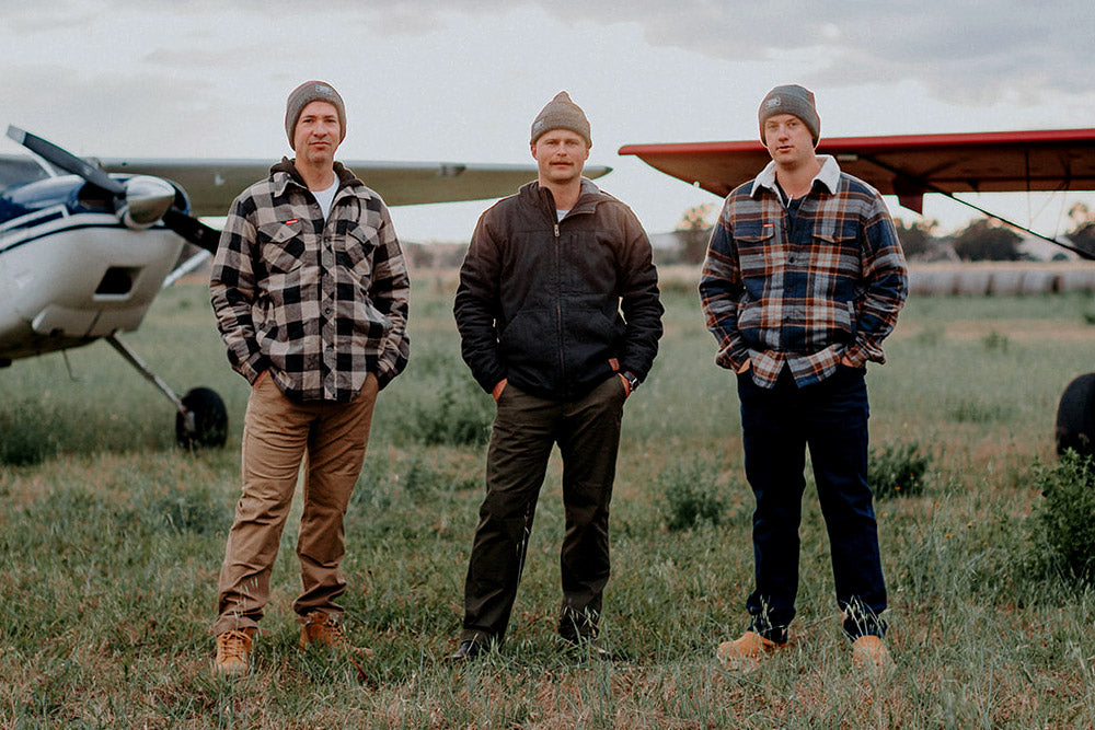 Three men standing in a grassy field with small airplanes in the background.