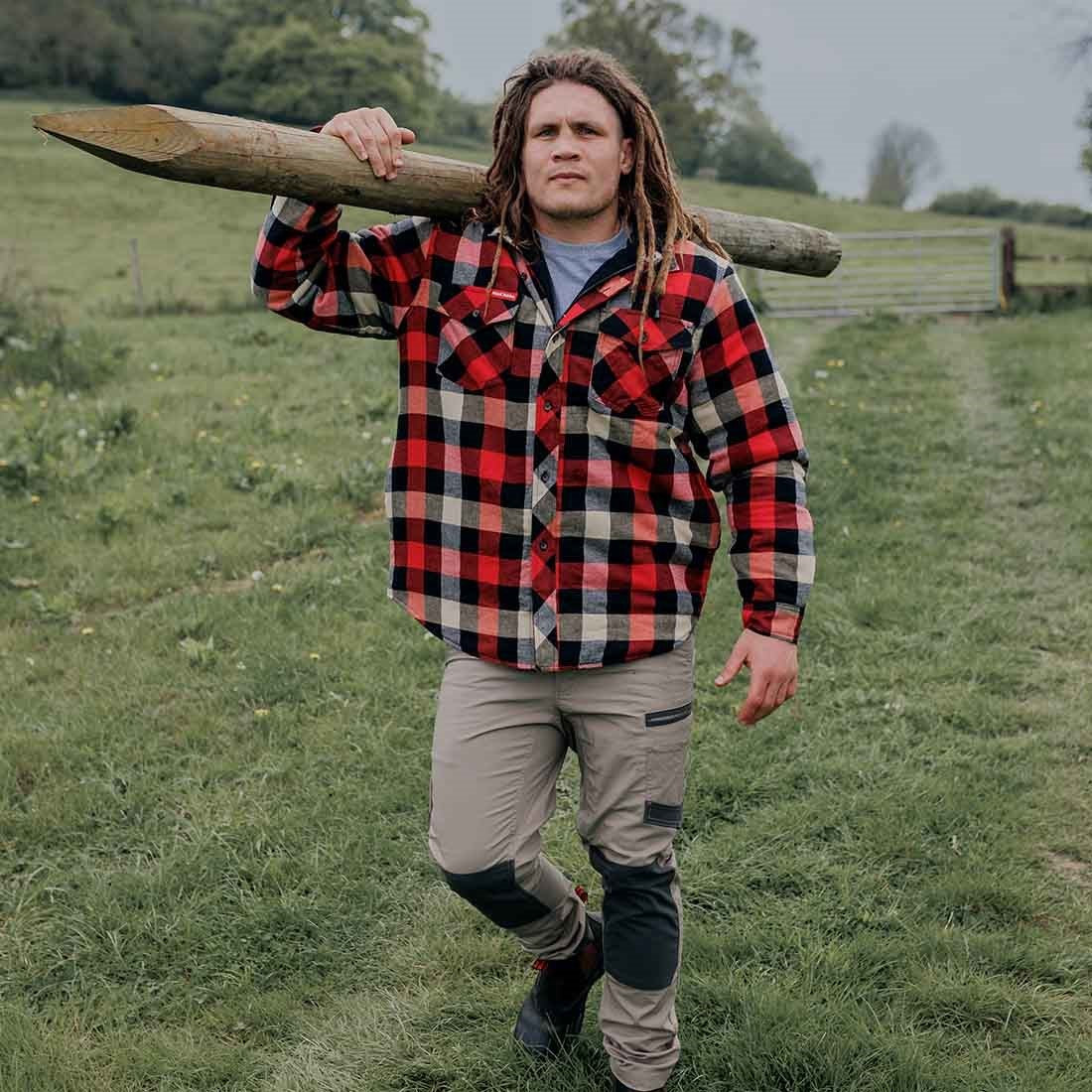 Person wearing a red and black checkered jacket carrying a log on a grassy field